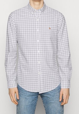 Mężczyzna ubrany w białą koszulę zapinaną na guziki z czarnym wzorem w kratę i haftowanym logo na piersi, w połączeniu z jasnoniebieskimi dżinsami.