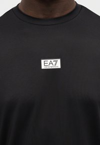 Μαύρο μπλουζάκι με στρογγυλή λαιμόκοψη από λείο, ελαστικό ύφασμα, με ορθογώνιο λευκό ετικέτα λογότυπου που φέρει το κείμενο "EA7 EMPORIO ARMANI" στο μπροστινό μέρος.
