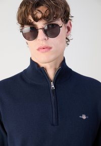 Pulover bleumarin cu guler înalt și închidere cu jumătate de fermoar. Fabricat dintr-un material moale, prezintă un logo mic pe piept și ochelari de soare rotunzi de culoare închisă.