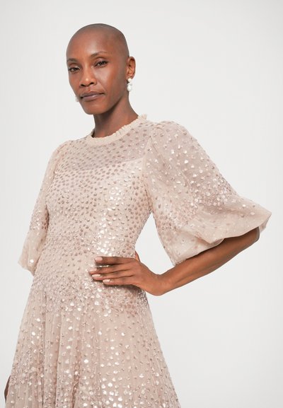 Beżowa sukienka z teksturowanej tkaniny, ozdobiona błyszczącymi cekinami. Ma krótkie bufiaste rękawy i falbaniaste wykończenie dekoltu.