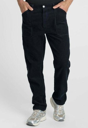 Zwarte corduroy broek gedragen door een persoon met handen in de zakken, gecombineerd met witte en grijze sportschoenen.