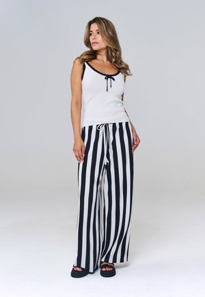 Žena s dlhými vlasmi, ktorá má na sebe biely tielko bez rukávov s čiernou lemovkou a čierno-biele vertikálne pruhované široké nohavice a čierne sandále.