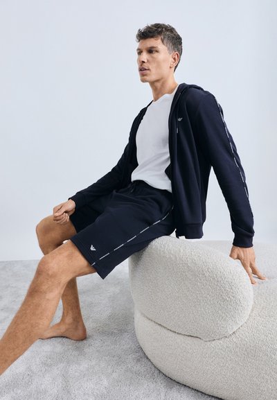 Sweat à capuche zippé marine avec logo blanc, assorti à un short marine avec des bandes latérales. Assis sur une chaise ronde texturée de couleur claire.