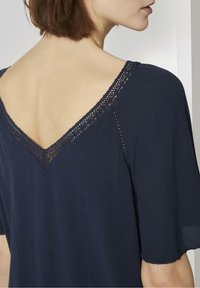 Donna che indossa una maglia blu navy con bordo in pizzo lungo il profondo scollo a V sulla schiena, che mostra la parte superiore della schiena scoperta e parte del volto di profilo.