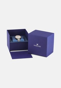 Reloj Swarovski en una caja morada con un interior de satén azul. El reloj presenta una cara redonda con un acabado brillante y una pulsera de metal.