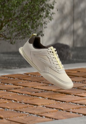 Beiger und brauner Freizeitsneaker, der über einem Boden mit Holz- und Kieselmuster schwebt, mit einer Betonwand und grüner Vegetation im Hintergrund.