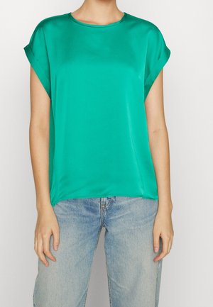 Woman wearing a short-sleeve teal blouse with rolled cuffs and light blue jeans, standing against a plain white background.