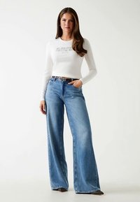 Top blanco de manga larga con detalle de texto; vaqueros de tiro alto y pierna ancha en azul claro; cinturón marrón; tela suave con un ajuste casual.