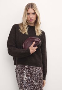 Barna kötött pulóver hosszú ujjal, flitteres szoknyával párosítva, burgundi színű clutch-táskával a kezében. Érezhető textúrájú anyagok és finom fény csillanása látható.