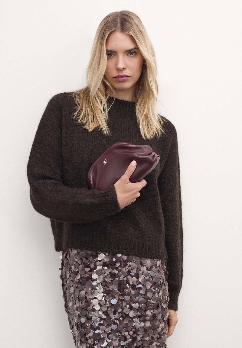 Barna kötött pulóver hosszú ujjal, flitteres szoknyával párosítva, burgundi színű clutch-táskával a kezében. Érezhető textúrájú anyagok és finom fény csillanása látható.