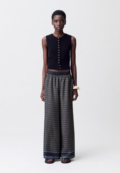 Una modelo se mantiene erguida, vistiendo una blusa negra sin mangas con botones dorados, combinada con pantalones anchos de estampado y sandalias sencillas.