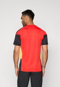 Raudona ir juoda sportinė marškinė su geltonais akcentais. Trumpomis rankovėmis, apvaliu iškirpimu, lygiu paviršiumi ir kontrastingu rašto dizainu.