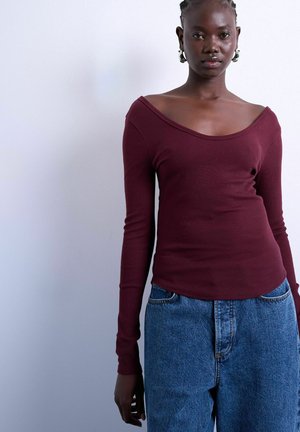 Nuori henkilö, jolla on tumma iho ja punotut hiukset, pukeutuneena viininpunaiseen olkapäät paljastavaan pitkähihaiseen paita ja sinisiin farkkuihin, valkoista taustaa vasten.