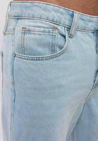 Jeans de mezclilla azul claro con un diseño de cinco bolsillos, costuras naranjas en contraste y una textura suave, mostrados en un ángulo lateral.