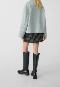 Veste grise en mélange de laine, mini-jupe à carreaux noire et hautes bottes en cuir noir avec accents dorés et fermetures éclair latérales.