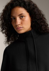 Čierny sveter s vysokým golierom z textúrovanej tkaniny, s uvoľneným strihom a šnúrkami, na neutrálnom pozadí.