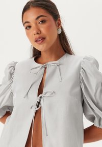 Blusa grigia chiaro con maniche corte a sbuffo, chiusura a fiocco al colletto, texture liscia e leggera lucentezza. Nessun motivo o componente visibile.
