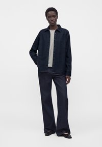 Femme vêtue d'une veste et d'un pantalon en jean foncé sur une chemise gris clair, debout avec une main dans la poche sur un fond uni.