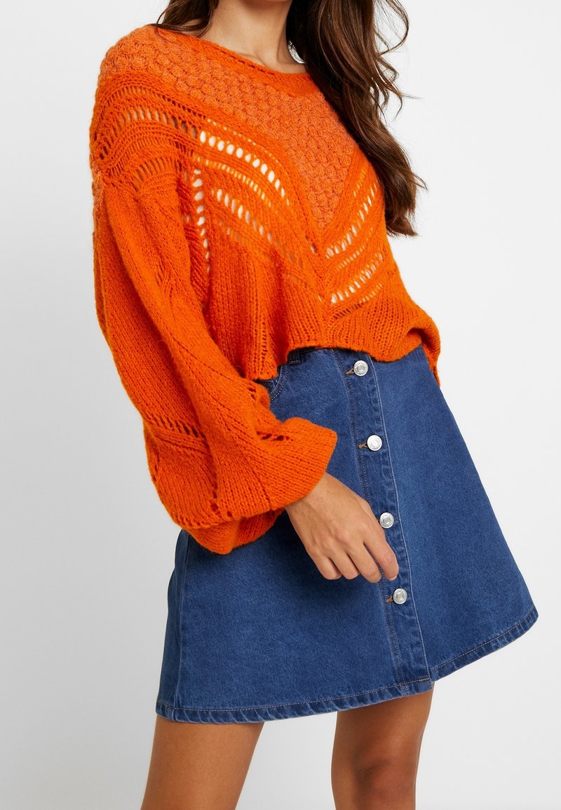 Jasnopomarańczowy, dziergany oversize sweter z otwartym wzorem ściegu i bufkami na rękawach, zestawiony z niebieską spódnicą dżinsową zapinaną na guziki z przodu.