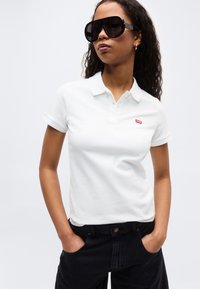 Γυναίκα που φοράει μεγάλα μαύρα γυαλιά ηλίου, άσπρο πολο με κόκκινο λογότυπο και μαύρα τζιν με τα χέρια στις τσέπες.