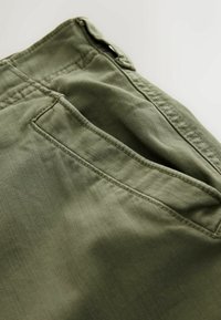 Gros plan d’un tissu vert olive avec une poche latérale cousue et des détails de coutures renforcées.