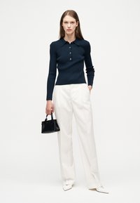 Γυναίκα που φοράει μπλε σκούρη πλεκτή μπλούζα με μακριά μανίκια, λευκά φαρδιά παντελόνια, λευκά μυτερά παπούτσια, κρατώντας μια μικρή μαύρη τσάντα μπροστά σε λευκό φόντο.