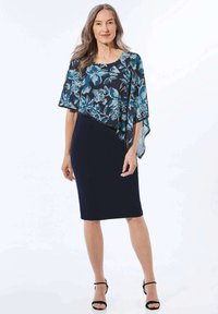 Blumige, durchscheinende Cape-Überlagerung in Blau und Türkis mit einem dunklen, taillierten Kleid darunter. Offene gesäumte Kanten. Strappy schwarze Sandalen runden das Outfit ab.
