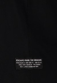 Schwarzer Stoff mit glatter Textur zeigt eine genähte Naht und ein Markenetikett, das weißen Text auf einem rechteckigen Patch anzeigt.