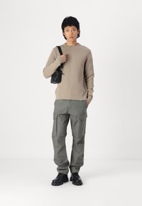 Beige ribbed knit sweater, gray cargo pants with pockets, black shoulder bag, and black boots. Model stands against a white background.