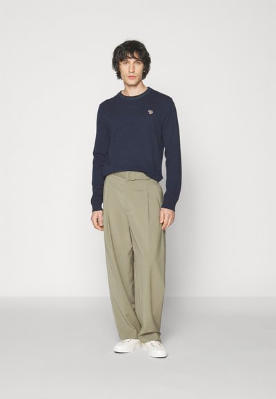 Pull en tricot marine à col rond, pantalon beige plissé et baskets blanches. Logo discret sur la poitrine du pull.