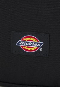 Črna tkaninska etiketa z logotipom blagovne znamke "Dickies", ki vključuje rumene in rdeče krožne elemente, obrobljene v modri barvi, z belim napisom.