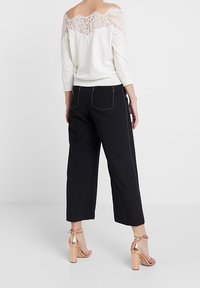 Top off-the-shoulder crema con dettagli in pizzo, abbinato a pantaloni neri a gamba larga e sandali con tacco a blocco in oro rosa.