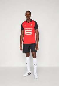 Červený dres s čiernymi rukávmi, na prednej strane biele číslo 5 a logo „SAMSIC“, doplnený o čierne šortky, biele ponožky a biele kopačky.