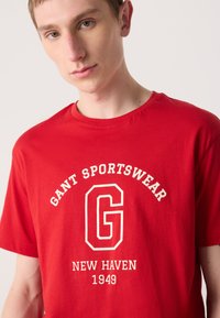 Rød bomulds t-shirt med rund hals, korte ærmer og hvid tekst, der står "GANT SPORTSWEAR NEW HAVEN 1949" med et emblematiske "G".