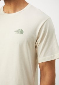 Beige cotton t-shirt with short sleeves and a crew neck. Features a green embroidered logo on the left chest area. Smooth texture.