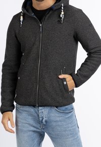 Grå zip-up hoodie med texturerad tyg, justerbar dragsko på huva och två framfickor. Matchat med ljusblå denimjeans.