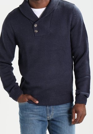 Ein Mann trägt einen dunkelblauen gestrickten Pullover mit Schalkragen und zwei Knöpfen, kombiniert mit hellblauen Jeans, und steht mit einer Hand in der Tasche.