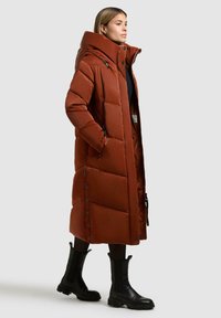 Cappotto lungo e gonfio di colore ruggine con grande cappuccio, bottoni a pressione e lacci neri. Indossato con stivaletti neri alla caviglia e presenta un design trapuntato.