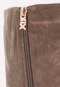 Brown suede boot featuring a gold-tone zipper and branded pull tab. The fabric has a soft texture with visible stitching.