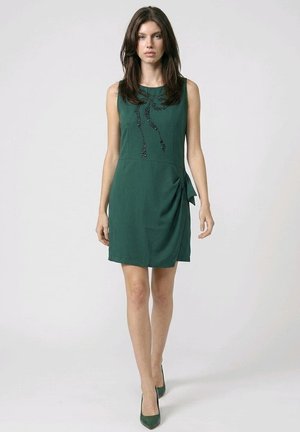 Mujer con vestido sin mangas de color verde oscuro hasta la rodilla con detalles de abalorios negros y tacones verdes a juego, de pie contra un fondo claro y liso.