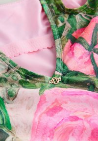 Nahaufnahme eines floralen Badeanzugs mit rosa und grünen Rosenmustern, die Nähte, Stofftextur und einen kleinen goldfarbenen dekorativen Anhänger am Ausschnitt hervorhebend.