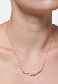 Collana in metallo dorato con un design curvato e ondulato. Catenina sottile con un pendente texturizzato, adagiata su un collo nudo.