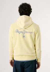 Felpa gialla con cappuccio regolabile, caratterizzata da un logo blu "Pepe Jeans" sul retro. Tessuto morbido con maniche lunghe e polsini a costine.