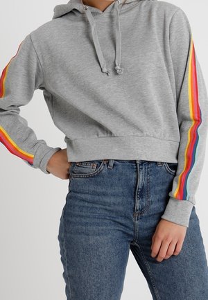 Personne portant un sweat à capuche court gris avec des bandes arc-en-ciel sur les manches et un jean bleu taille haute, posant avec une main sur la hanche.
