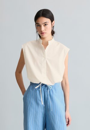 Młoda kobieta o ciemnych włosach nosi bezrękawnik z białymi guzikami oraz niebieskie, wysokowalne spodnie w paski ze sznurkiem, stoi na jasnoszarym tle.