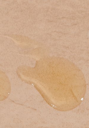 Beiger Stoff mit glatter Textur, der zwei klare Tropfen Flüssigkeit zeigt, einen größeren und unregelmäßig geformten, der auf der Oberfläche ruht.