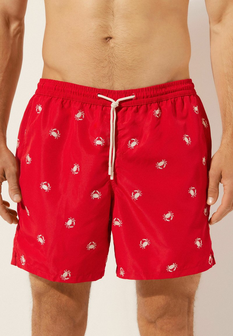 Shorts de bain rouges avec des motifs de crabes blancs, taille élastique et cordon de serrage. Matériau léger, texture lisse et longueur au-dessus du genou.