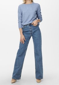 Femme portant un pull bleu clair, un jean bleu taille haute à jambes droites, et des talons beiges à bout pointu, debout devant un fond blanc uni.