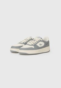 Sapatilhas cinzentas e off-white com um cabedal de couro suave, detalhes texturizados, biqueira redonda, atacadores planos e uma sola de borracha grossa. Logótipo da Lacoste presente.