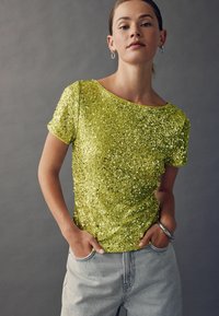 Glittrig gul paljettröja med rund halsringning, korta ärmar och avslappnad passform, ihop med ljusblå hög midja jeans.